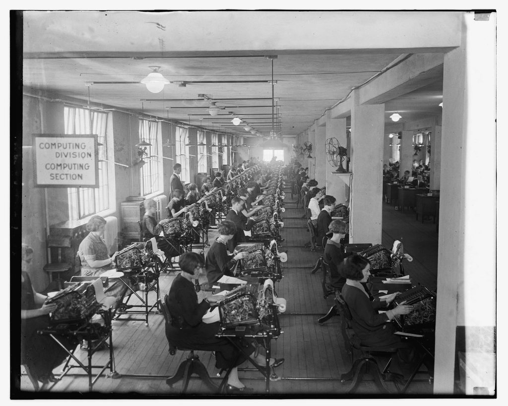 A black-and-white photograph of a long 1920s office, the Computing Division, with rows of clerks working at mechanical calculating machines.