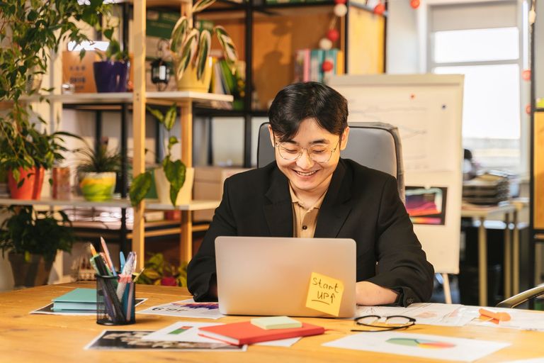 A young professional smiles while working at a laptop in a bright office.